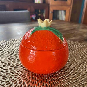 May include: A round, bright orange ceramic lidded jar shaped like a piece of citrus fruit. The lid features a textured, bumpy surface with a green leaf and a small, pale yellow flower bud at the top center, mimicking a real orange.
