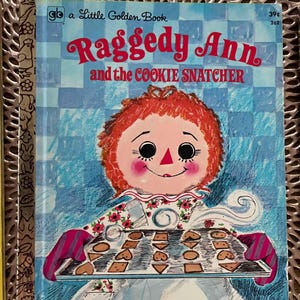 May include: A Little Golden Book titled "Raggedy Ann and the Cookie Snatcher." The cover features a Raggedy Ann illustration holding a tray of cookies. The background is a blue and white checkered pattern.