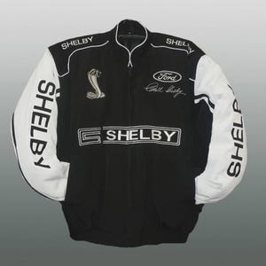 Op de afbeelding: Zwart-witte Shelby racejas. De jas heeft witte mouwen met verticaal gedrukt "SHELBY". De voorkant toont "SHELBY" en een geborduurd cobra-logo, plus de tekst "Ford" en "Carroll Shelby". Een klassieke jas met racethema.