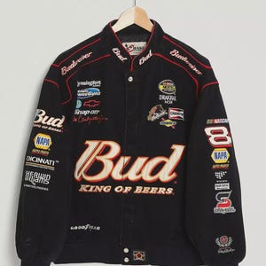 Puede incluir: Chaqueta de carreras negra con múltiples parches de patrocinadores, incluyendo "Bud", "NASCAR" y "Goodyear". La chaqueta presenta detalles bordados en rojo y blanco y una parte delantera abotonada. El texto "King of Beers" se muestra de forma destacada.