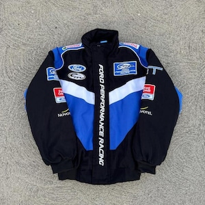 Op de afbeelding: Zwarte, blauwe en witte Ford Performance Racing jas. De jas heeft een zwarte body met blauwe en witte accenten en meerdere Ford-logo's. De tekst "FORD PERFORMANCE RACING" is verticaal op de voorkant gedrukt.