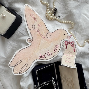 May include: A watercolor dove-shaped save-the-date card with the names "Celine & Gerard" and the date "16th of May 2026, Paris, France." The card is accompanied by a pearl necklace and jewelry boxes.