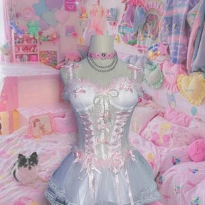 Puede incluir: Un vestido de tul y encaje blanco y rosa con un corpiño de corsé y un collar de corazón. El vestido se muestra en una habitación con un esquema de color rosa y blanco.