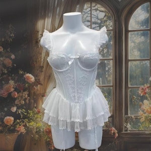 Puede incluir: Lencería blanca de encaje con un corpiño de corsé, falda con volantes y ligas. La lencería está en un maniquí blanco frente a una ventana.