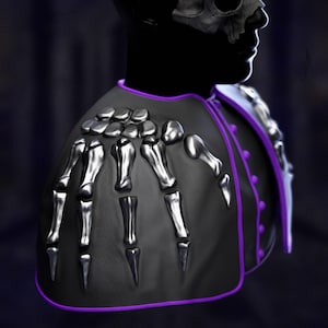 May include: A black shoulder cape with a skeleton hand design. The cape features silver bone accents and a purple trim. The design is set against a dark background, creating a gothic or fantasy aesthetic.