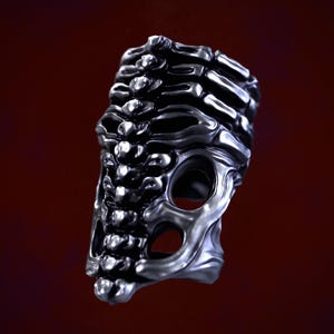 May include: A silver ring with a skeletal design. The ring is made of metal and has a textured surface. The ring is designed to look like a rib cage.