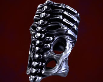 May include: A silver ring with a skeletal design. The ring is made of metal and has a textured surface. The ring is designed to look like a rib cage.