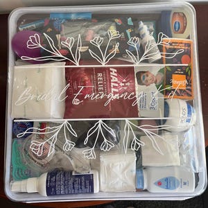 May include: A clear plastic organizer case filled with various personal care items, including Halls cough drops, Kleenex, Vaseline, and Johnson's baby powder. The lid features white floral line art and the text Bridal Emergency Kit in script.