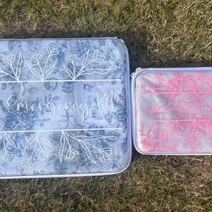 Puede incluir: Dos estuches rectangulares con cremallera y estampado floral. El estuche m&aacute;s grande presenta l&iacute;neas florales azules y el texto "Bridal Emergency Kit" en blanco. El m&aacute;s peque&ntilde;o presenta l&iacute;neas florales rosas y el mismo texto en blanco.