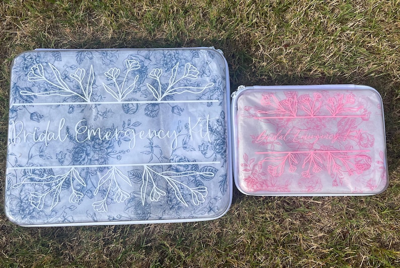 Puede incluir: Dos estuches rectangulares con cremallera y estampado floral. El estuche m&aacute;s grande presenta l&iacute;neas florales azules y el texto "Bridal Emergency Kit" en blanco. El m&aacute;s peque&ntilde;o presenta l&iacute;neas florales rosas y el mismo texto en blanco.