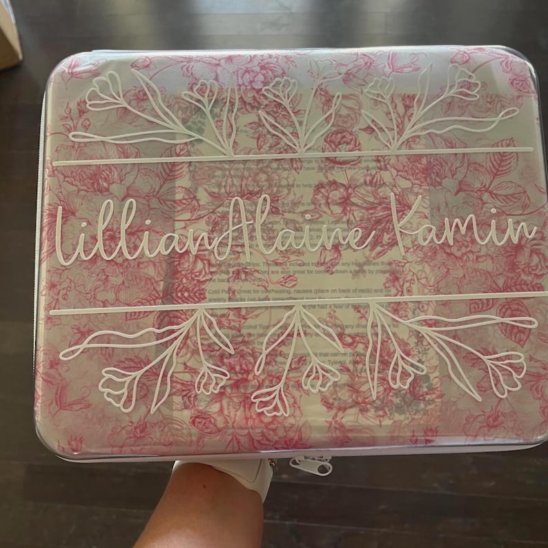 Puede incluir: Un estuche de almacenamiento rectangular transparente con un patr&oacute;n floral rosa y el texto en letra cursiva blanca Lillian Alaine Kamin centrado entre dos l&iacute;neas horizontales con ilustraciones bot&aacute;nicas. Tiene un cierre de cremallera.