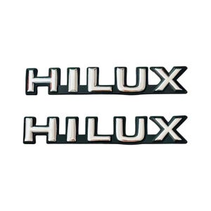Peut inclure: Deux emblèmes "HILUX" argentés sur fond noir.