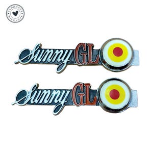 Puede incluir: Dos emblemas de coche plateados con las palabras "Sunny GL" en letras rojas y negras. Cada emblema tiene un diseño redondo en forma de diana, blanco, amarillo y rojo.