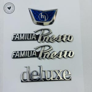 May include: A set of four silver car badges. The top badge is a blue and silver crest with the letter "m" in the centre. The next two badges are silver and black and read "FAMILIA PRESTO". The bottom badge is silver and reads "deluxe".