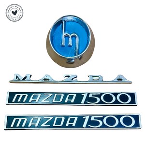 Könnte beinhalten: Ein Satz von drei silbernen Mazda-Autoemblemen. Das erste Emblem ist ein rundes, blaues Emblem mit dem Mazda-Logo. Das zweite Emblem ist eine silberne Leiste mit dem Wort "MAZDA" in erhabenen Buchstaben. Das dritte und vierte Emblem sind schwarze rechteckige Embleme mit den Worten "MAZDA 1500" in weißen Buchstaben.