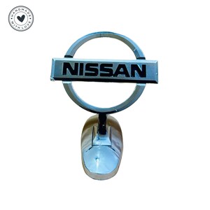 Puede incluir: Un emblema de coche plateado con un logotipo NISSAN negro en un c&iacute;rculo. El emblema est&aacute; unido a una base plateada.