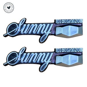 Könnte beinhalten: Zwei silberne und schwarze Autoembleme mit dem Wort "Sunny" in einer kursiven Schriftart und dem Wort "Deluxe" in einer Blockschrift.