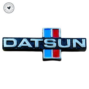 May include: A silver Datsun emblem with black lettering and a red, white, and blue stripe design.