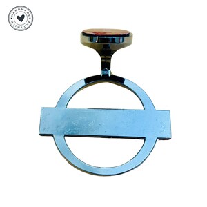 Puede incluir: Un emblema de coche de metal plateado con un c&iacute;rculo y una barra horizontal. El emblema tiene un dise&ntilde;o rojo y blanco en la parte superior.