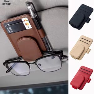 Könnte beinhalten: Ein Auto-Sonnenblenden-Organizer aus braunem Leder mit zwei Kartenfächern, einem Stiftehalter und einem Magnetclip für Brillen. Der Organizer ist an einer grauen Sonnenblende befestigt. Weitere Farbvarianten in Schwarz, Beige und Rot.