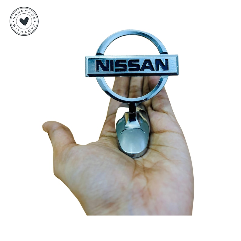 Puede incluir: Un emblema de coche plateado con un logotipo NISSAN negro dentro de un c&iacute;rculo. El emblema est&aacute; unido a una base plateada.