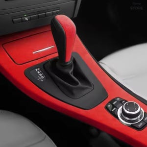 Peut inclure: Une console centrale de voiture avec un panneau de garniture rouge vif, un pommeau de levier de vitesse noir avec un insert rouge sur le dessus et un soufflet de levier de vitesse en cuir noir. La console inclut une molette et un indicateur P, R, N, D.