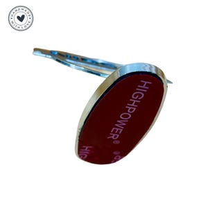 Puede incluir: Un soporte para tel&eacute;fono de forma ovalada de color plateado con un respaldo adhesivo rojo. El soporte tiene el texto "HIGHPOWER&reg;" impreso en el respaldo adhesivo.