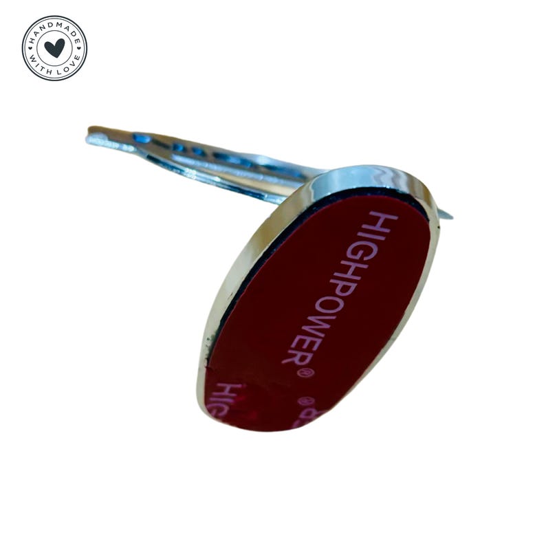 Puede incluir: Un soporte para tel&eacute;fono de forma ovalada de color plateado con un respaldo adhesivo rojo. El soporte tiene el texto "HIGHPOWER&reg;" impreso en el respaldo adhesivo.