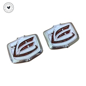 Puede incluir: Dos gemelos de metal plateado con un diseño de esmalte rojo y blanco. El diseño presenta una letra "E" estilizada con una estrella a cada lado.