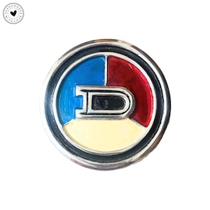 May include: A round, silver-toned emblem with a segmented design. The top section is divided into blue and red, with a yellow lower section. A black and silver letter "D" is in the center. The emblem is surrounded by a black border.