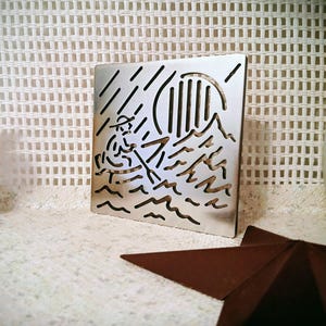 May include: A square, silver-colored metal art piece featuring a cutout design of a person rowing a boat on wavy water, with a sun and rain in the background. The piece is standing upright on a white surface.