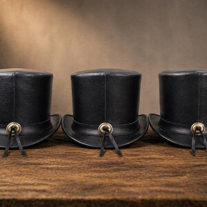 Puede incluir: Tres sombreros de copa de cuero negro sobre una superficie de madera. Cada sombrero tiene una copa redondeada, un ala ancha y un bot&oacute;n dorado con lazos de cuero negro. Dise&ntilde;o cl&aacute;sico.