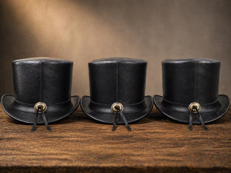 Puede incluir: Tres sombreros de copa de cuero negro sobre una superficie de madera. Cada sombrero tiene una copa redondeada, un ala ancha y un bot&oacute;n dorado con lazos de cuero negro. Dise&ntilde;o cl&aacute;sico.