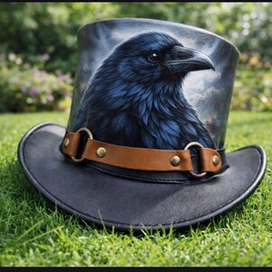 Puede incluir: Un sombrero de copa de cuero negro con una pintura detallada de un cuervo en la corona. El cuervo est&aacute; representado con plumas azul oscuro y un pico negro. Una banda de cuero marr&oacute;n con anillos y tachuelas de color lat&oacute;n rodea el sombrero.