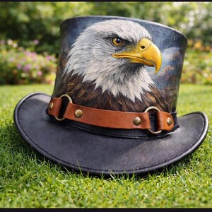 Puede incluir: Un sombrero de copa negro con una pintura detallada de la cabeza de un &aacute;guila calva. El sombrero presenta una banda de cuero marr&oacute;n con anillos y tachuelas de metal color lat&oacute;n. Las plumas del &aacute;guila son blancas y marrones, con un pico amarillo.