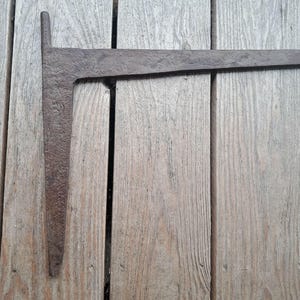 May include: A dark brown, weathered iron L-shaped tool with a textured, pitted surface. The object features a long horizontal arm and a shorter, tapered vertical arm, showing signs of age and oxidation across its entire metal construction.