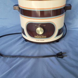 May include: Cream-colored Rival Crock-Pot slow cooker with brown and blue accents. It has a glass lid and a control dial with settings for off, low, and high. The power cord is plugged in.