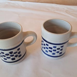 May include: Two ceramic mugs with a light gray glaze and a dark blue painted pattern. Each mug features a cylindrical body, a sturdy side handle, and a decorative band of blue squares and horizontal lines encircling the lower portion of the exterior.