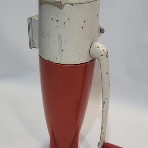 May include: A vintage manual food mill featuring a cream-colored cylindrical top housing with a red circular label on the lid, attached to a tapered red plastic base with three fins. A white metal hand crank with a red knob extends from the side.