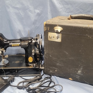 May include: A vintage Singer sewing machine in black with gold accents, a foot pedal, and a black carrying case. The machine has the Singer logo and is set against a blue backdrop. The case has metal clasps.