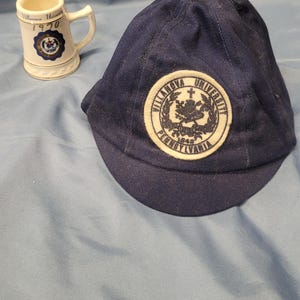 May include: A navy blue cap with a circular patch featuring the words "Villanova University Pennsylvania" and a crest. Beside it is a small white mug with the same crest and the words "Villanova University 1970".