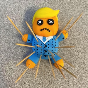 May include: A novelty voodoo doll with yellow hair, an orange face, and a blue suit. The doll has black cross eyes and a stitched frown. Wooden toothpicks are inserted into the doll's body, arms, and legs.