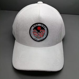 Op de afbeelding: Witte baseballpet met een gebogen klep. De pet heeft een cirkelvormige patch met de tekst "GIBO" en "NEW YAPRK" rond een ontwerp van een rode zon en blauwe en rode golven.