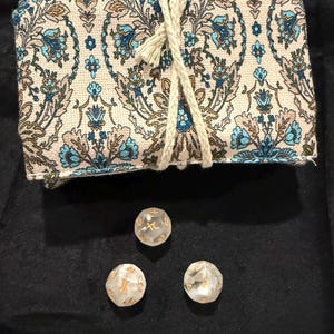 May include: A beige and blue floral patterned dice bag tied with a cream-coloured rope, with three clear dice with gold lettering. The dice bag is rectangular and folded over. The dice are translucent and have gold markings.