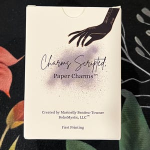 May include: A white box with the text "Charms Scripted, Paper Charms" and a hand illustration. The box is labeled "Created by Marinelly Benitez-Towner BohoMystic, LLC" and "First Printing".