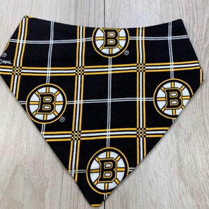 May include: Black and gold plaid dog bandana featuring the Boston Bruins logo. The bandana has a repeating pattern of the team's logo within a grid of white and gold lines. The NHL logo is visible in the corner.