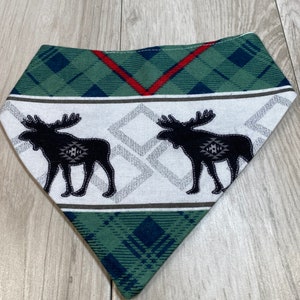 May include: A green, white and black patterned bandana with two moose silhouettes. The bandana has a red and green plaid border.