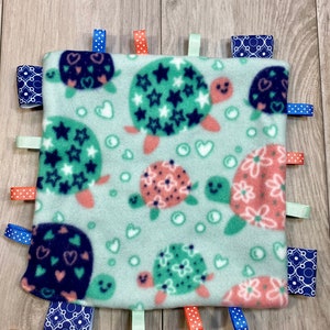 May include: A soft, green baby blanket with a turtle pattern. The blanket has multiple colorful ribbons attached to the edges.