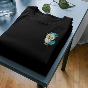 以下が含まれることがあります： 前面に青と白の漫画の鳥が刺繍された黒いTシャツ