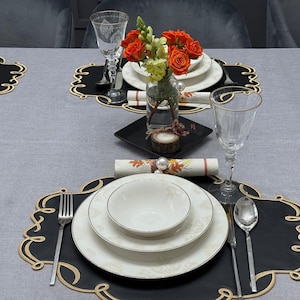 May include: A formal dining table setting with white plates, bowls, and silverware. Black placemats with gold trim are under each setting. A vase of orange roses and greenery sits in the center. Wine glasses and a rolled napkin complete the elegant arrangement.
